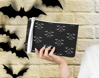May include: A black zippered pouch with a white bat pattern. The pouch is held in a hand against a brick wall with a bat silhouette pattern.