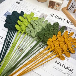 May include: A collection of crocheted leaf stems in various shades of green and yellow. The stems are arranged on a newspaper background, with the leaves in a variety of colors, including dark green, light green, and yellow.