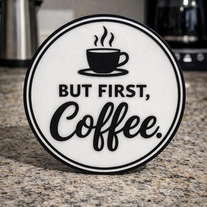 Peut inclure: Un dessous de verre rond blanc avec une bordure noire. Il présente une icône de tasse à café noire et les mots "BUT FIRST, Coffee." en noir. Le dessous de verre est posé sur un comptoir moucheté.
