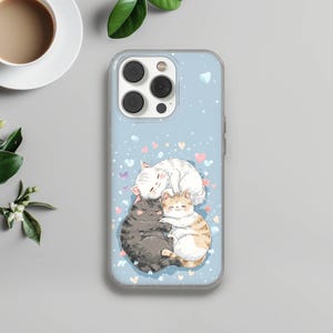 May include: A light blue phone case featuring a watercolor illustration of four cuddling cats. The cats are in shades of gray, white, and orange, with hearts and dots scattered around them. The case has a clear border and a camera cutout.