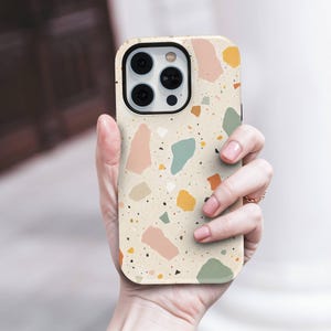 Puede incluir: Una funda para teléfono con fondo color crema y un patrón de terrazo multicolor. El diseño incluye formas irregulares dispersas en tonos rosa empolvado, verde salvia, amarillo mostaza y marrón oscuro, junto con pequeñas motas negras.