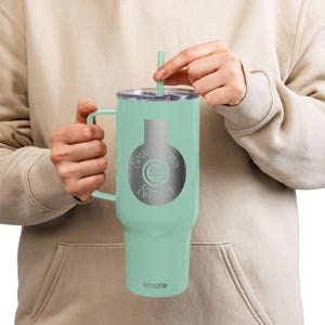 May include: A mint green insulated tumbler with a handle and a lid, featuring a silver logo that reads "Small Town Steam". The tumbler includes a matching green straw. The brand name "koozie" is printed at the bottom.
