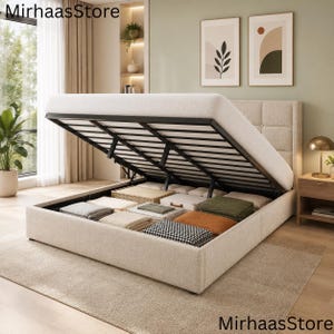 Op de afbeelding: Een beige gestoffeerd opbergbed met een gecapitonneerd hoofdeinde en een hefmechanisme dat een zwart metalen frame met houten latten onthult. De interne opbergruimte bevat diverse gevouwen dekens en textiel in neutrale en gedempte tinten.