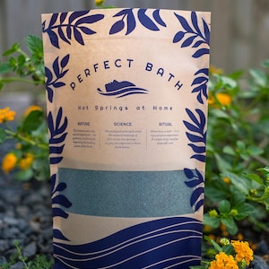 May include: A tan-colored stand-up pouch with dark blue botanical illustrations and text that reads PERFECT BATH, Hot Springs at Home, Nature, Science, and Ritual. The pouch features a clear horizontal window revealing a greyish-green powder inside.