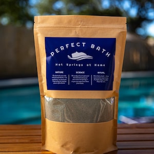 May include: A kraft paper bag with a blue label that reads "PERFECT BATH Hot Springs at Home." The bag contains a dark gray powder. The text on the label describes the product's nature, science, and ritual.