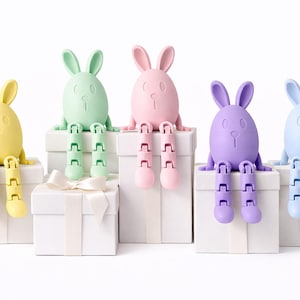 May include: Five pastel-colored bunny figurines with articulated legs sit atop white gift boxes. The bunnies are yellow, green, pink, purple, and blue. The image is set against a white background, with one box tied with a white ribbon.