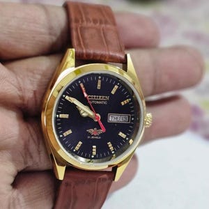 Pode incluir: Um relógio automático Citizen dourado com mostrador azul escuro e pulseira de couro castanho. O mostrador do relógio exibe o dia e a data, com ponteiros e marcadores dourados. A pulseira é de uma rica cor castanha.