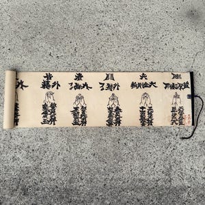 May include: A beige paper scroll featuring black ink calligraphy and illustrations of hands in a prayer position. The scroll displays multiple columns of traditional characters and symbols, with a wooden rod at the left end and a black string attached.