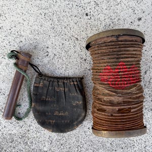 Könnte beinhalten: Eine zylindrische, zusammenklappbare braune Papierlaterne mit rotem Blumenemblem, ein kleiner schwarzer Stoffbeutel mit wiederkehrendem goldenem Muster und ein dunkelbrauner Holzgriff an einer grünen Kordel, angeordnet auf einer grauen Oberfläche.
