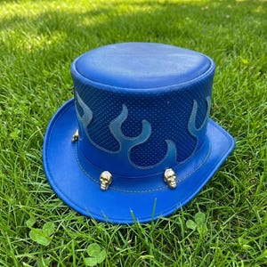 May include: A striking blue top hat with flame detailing and gold skull studs. The hat has a wide brim and a textured crown, set on a bed of green grass.
