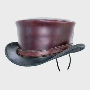 May include: A burgundy leather top hat with a black brim and a brown leather band. The hat has a classic tall crown and a slightly curved brim. Two black strings hang from the brim.