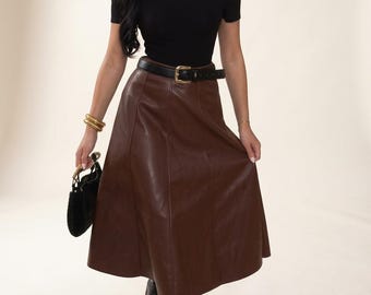 Könnte beinhalten: Ein schwarzes schulterfreies Oberteil kombiniert mit einem braunen wadenlangen Rock mit vertikalen Nahtdetails. Das Outfit umfasst einen schwarzen Gürtel mit goldfarbener Schnalle, eine schwarze Handtasche mit goldenen Beschlägen und schwarze Stiefel.