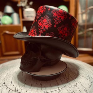 Könnte beinhalten: Ein schwarzer Zylinder mit einem sich wiederholenden roten Damastmuster und Totenkopfmotiven. Der Hut wird auf einem schwarzen, totenkopfförmigen Ständer präsentiert. Das Design umfasst komplizierte Schnörkel um jeden Totenkopf, die einen starken Kontrast zum schwarzen Hintergrund der Krone und Krempe des Hutes bilden.