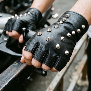 Peut inclure: Une paire de gants noirs sans doigts avec une surface texturée et de multiples clous coniques en métal argenté disposés sur le dos de la main et le poignet. Les gants sont conçus avec des bouts de doigts ouverts et un poignet court.