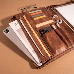 May include: A brown leather portfolio case with multiple compartments, including card slots, pen holders, and a phone pocket. The case is open, revealing a tablet, notebook, and various accessories. A pair of eyeglasses rests on top.