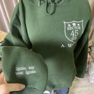 Peut inclure: Un sweat &agrave; capuche vert avec du texte brod&eacute; blanc sur la manche qui dit "Ignite, my love. Ignite." Le sweat &agrave; capuche pr&eacute;sente &eacute;galement un &eacute;cusson blanc avec les mots "Sector 45 CCR" imprim&eacute;s sur le devant.