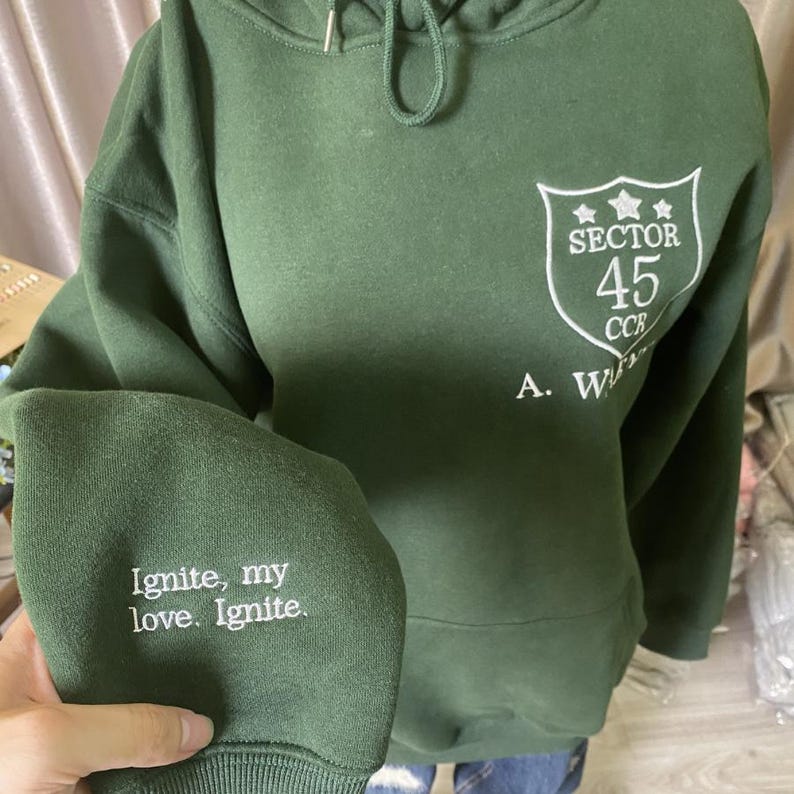 Peut inclure: Un sweat &agrave; capuche vert avec du texte brod&eacute; blanc sur la manche qui dit "Ignite, my love. Ignite." Le sweat &agrave; capuche pr&eacute;sente &eacute;galement un &eacute;cusson blanc avec les mots "Sector 45 CCR" imprim&eacute;s sur le devant.