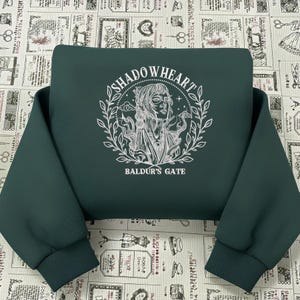 Può includere: Felpa verde scuro con grafica bianca di un volto femminile all'interno di un disegno circolare. Il disegno include il testo "SHADOWHEART" sopra l'immagine e "BALDUR'S GATE" sotto. La felpa ha polsini e orlo a coste.