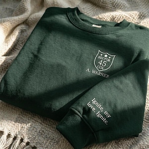 Peut inclure: Un sweat-shirt vert fonc&eacute; avec du texte et des graphiques blancs. La poitrine pr&eacute;sente un &eacute;cusson avec "SECTOR 45 CCR" et "A. WARNER". La manche indique "Ignite, my love. Ignite."