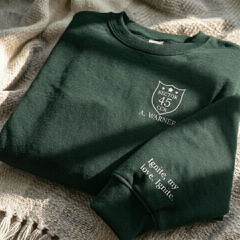 Peut inclure: Un sweat-shirt vert fonc&eacute; avec du texte et des graphiques blancs. La poitrine pr&eacute;sente un &eacute;cusson avec "SECTOR 45 CCR" et "A. WARNER". La manche indique "Ignite, my love. Ignite."