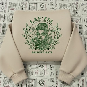 May include: A beige sweatshirt featuring a green graphic of a character with pointed ears, surrounded by a laurel wreath. The text "LAE'ZEL" is above the character, and "BALDUR'S GATE" is below. The sweatshirt has long sleeves.
