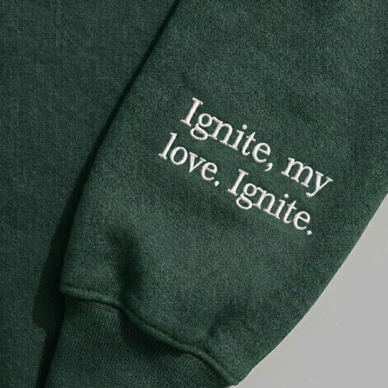 Peut inclure: Manche de sweat-shirt vert fonc&eacute; avec texte blanc brod&eacute; : "Ignite, my love. Ignite."