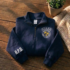 Może przedstawiać: Granatowa bluza z kołnierzem zapinanym na zamek 1/4. Z przodu znajduje się okrągła odznaka z napisem "S.T.A.R.S. Raccoon Police Dept." i dwiema żółtymi gwiazdami. Lewy rękaw wyświetla "R.P.D." w białych literach. Bluza leży na drewnianej powierzchni.