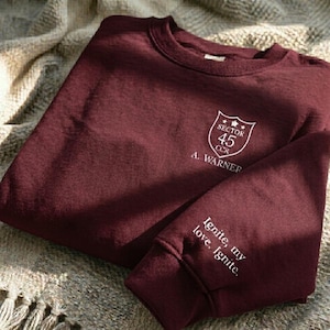 Peut inclure: Un sweat-shirt bordeaux avec du texte blanc et un &eacute;cusson. L'&eacute;cusson indique "Sector 45 CCR" et "A. Warner". La manche affiche le texte "Ignite, my love, Ignite."