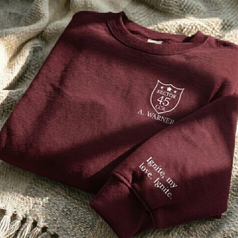 Peut inclure: Un sweat-shirt bordeaux avec du texte blanc et un &eacute;cusson. L'&eacute;cusson indique "Sector 45 CCR" et "A. Warner". La manche affiche le texte "Ignite, my love, Ignite."