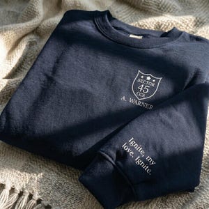 Peut inclure: Sweat-shirt bleu marine avec texte blanc et un &eacute;cusson. L'&eacute;cusson indique "Sector 45 CCR" et "A. Warner". La manche porte l'inscription "Ignite, my love, Ignite."