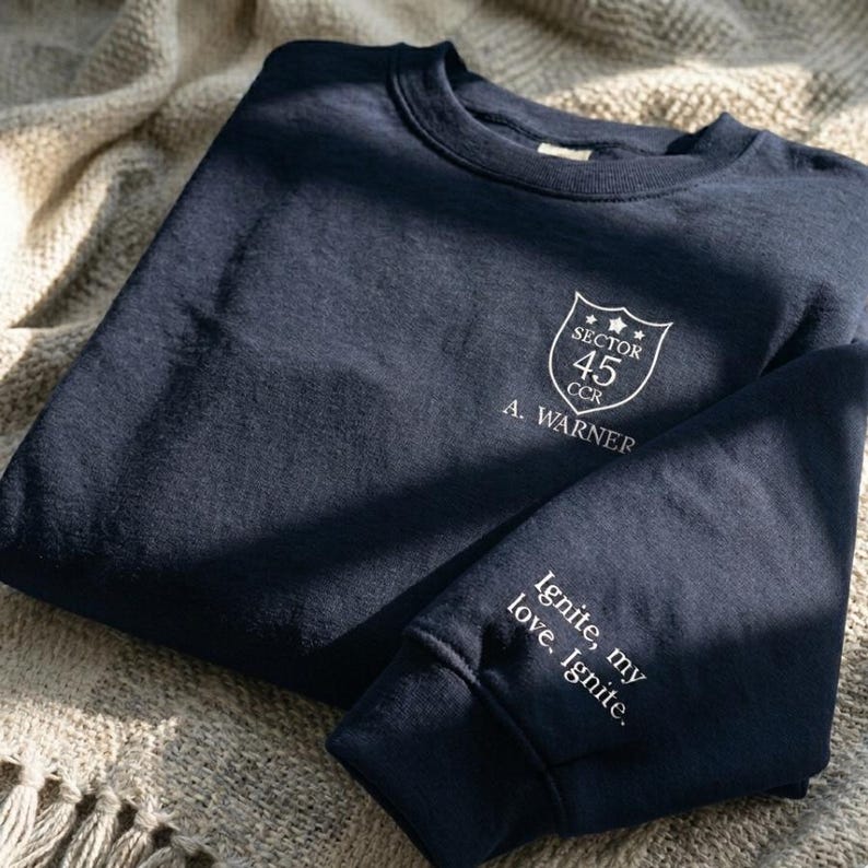 Peut inclure: Sweat-shirt bleu marine avec texte blanc et un &eacute;cusson. L'&eacute;cusson indique "Sector 45 CCR" et "A. Warner". La manche porte l'inscription "Ignite, my love, Ignite."