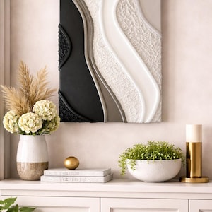 May include: Abstract wall art with a textured design in black, white, and gray. The artwork is mounted above a white cabinet, which holds a vase of flowers, books, a gold sphere, and a small plant in a white bowl. A gold and white lamp is also on the cabinet.