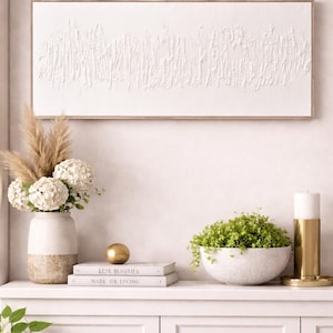 May include: A white textured abstract art piece in a wooden frame hangs above a white cabinet. The cabinet displays a vase with flowers, books, a gold sphere, a bowl of greenery, and a gold candle holder.