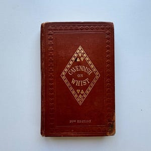 May include: A vintage book titled "Cavendish on Whist." The cover is a deep reddish-brown with gold lettering and diamond-shaped design. The text "20th Edition" is at the bottom. The book is in good condition.