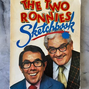 Op de afbeelding: Vintage boekcover "The Two Ronnies Sketchbook" met twee lachende mannen met een bril. De titel staat in rode en blauwe letters met een blauwe verfspat. De mannen dragen pakken en stropdassen.
