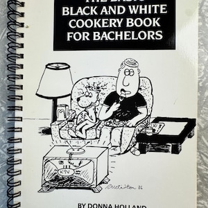 May include: A spiral-bound cookbook titled "THE BASIC BLACK AND WHITE COOKERY BOOK FOR BACHELORS." The cover features a black and white cartoon illustration of a man and dog watching TV. The book is by Donna Holland.