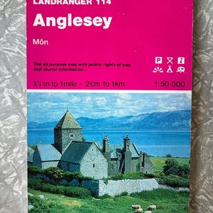 May include: A vintage Ordnance Survey map of Anglesey, Wales, with a bright pink cover. The map features a scenic illustration of Penmon Priory and includes text indicating a scale of 1½ inches to 1 mile.