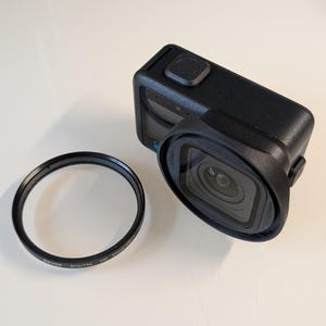 May include: A black action camera with a protective housing and a circular lens filter. The camera has a square lens and a button on top. The lens filter has text that reads "DIGITAL HD Close-Up".