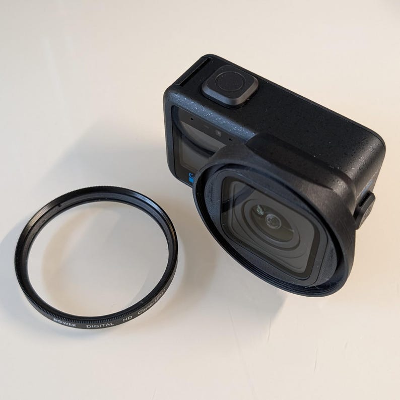 May include: A black action camera with a protective housing and a circular lens filter. The camera has a square lens and a button on top. The lens filter has text that reads "DIGITAL HD Close-Up".