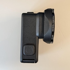 May include: A black action camera with a textured matte finish and a square button on the side. Attached to the front is a black circular lens filter housing with the text "ATONE FOCUSXTENDER 55mm" printed clearly along the outer edge.