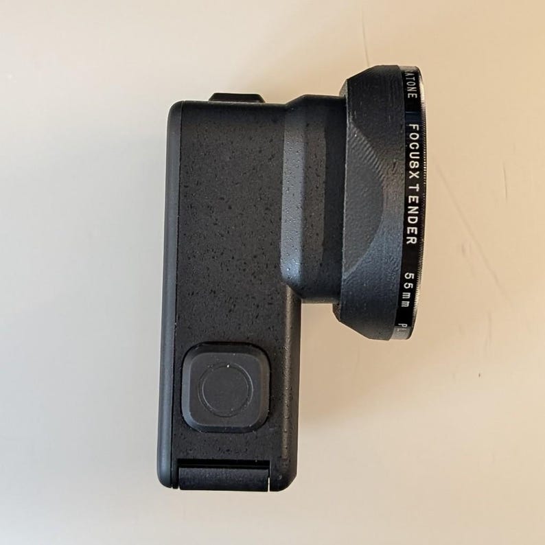 May include: A black action camera with a textured matte finish and a square button on the side. Attached to the front is a black circular lens filter housing with the text "ATONE FOCUSXTENDER 55mm" printed clearly along the outer edge.