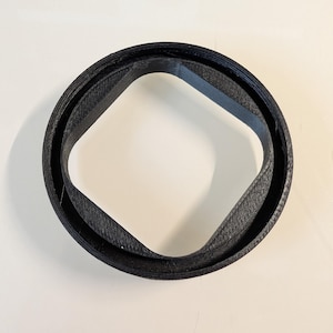 May include: A black circular object with a textured surface featuring a square-shaped inner opening with rounded corners. The item has a layered construction, showing visible horizontal lines along the outer rim and the inner square border.