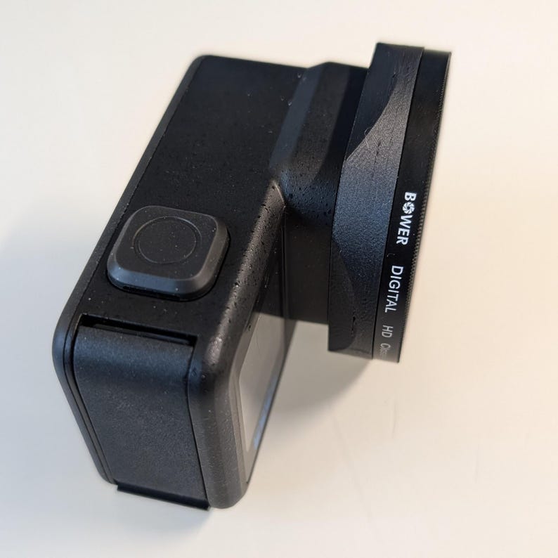 May include: A black digital camera with a lens attached. The camera has a rectangular shape with a prominent button on the top. The lens is labeled "Bower Digital HD Optics".