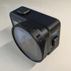 K&ouml;nnte beinhalten: Eine schwarze Action-Kamera mit einem Weitwinkelobjektiv, das von einem texturierten schwarzen Geh&auml;use gesch&uuml;tzt wird. An der Seite des Ger&auml;ts befinden sich der Schriftzug MISSION 1 PRO und ein Ein-/Ausschalter mit der Aufschrift MODE.