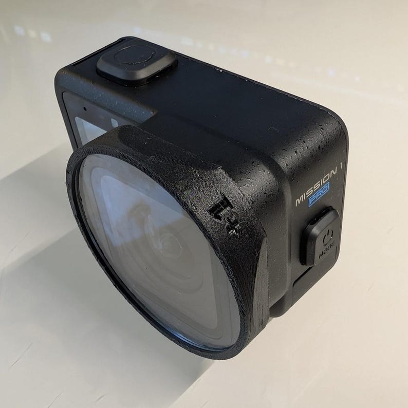 K&ouml;nnte beinhalten: Eine schwarze Action-Kamera mit einem Weitwinkelobjektiv, das von einem texturierten schwarzen Geh&auml;use gesch&uuml;tzt wird. An der Seite des Ger&auml;ts befinden sich der Schriftzug MISSION 1 PRO und ein Ein-/Ausschalter mit der Aufschrift MODE.