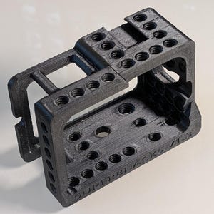 May include: A black 3D-printed camera cage featuring a rectangular frame with multiple threaded mounting holes on the top and side panels. The side of the frame is embossed with the text SUPERHACKS V1.3, and the structure has a textured finish.