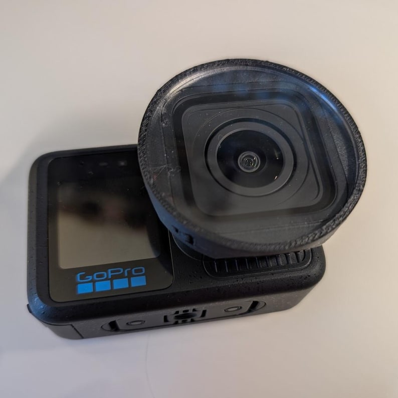 K&ouml;nnte beinhalten: Eine schwarze GoPro Action-Kamera mit einem Frontdisplay und dem blauen GoPro-Logo. Ein kreisf&ouml;rmiger schwarzer Objektivfilter oder ein Schutzgeh&auml;use ist &uuml;ber dem quadratischen Kameraobjektiv angebracht und ragt aus dem Geh&auml;use hervor.