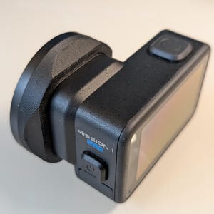 May include: A black action camera with a lens attachment. The camera has a rectangular body with a screen and a button on top. The text "MISSION 1 PRO" is visible on the side. The lens attachment is circular and black.