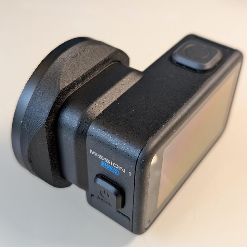 May include: A black action camera with a lens attachment. The camera has a rectangular body with a screen and a button on top. The text "MISSION 1 PRO" is visible on the side. The lens attachment is circular and black.