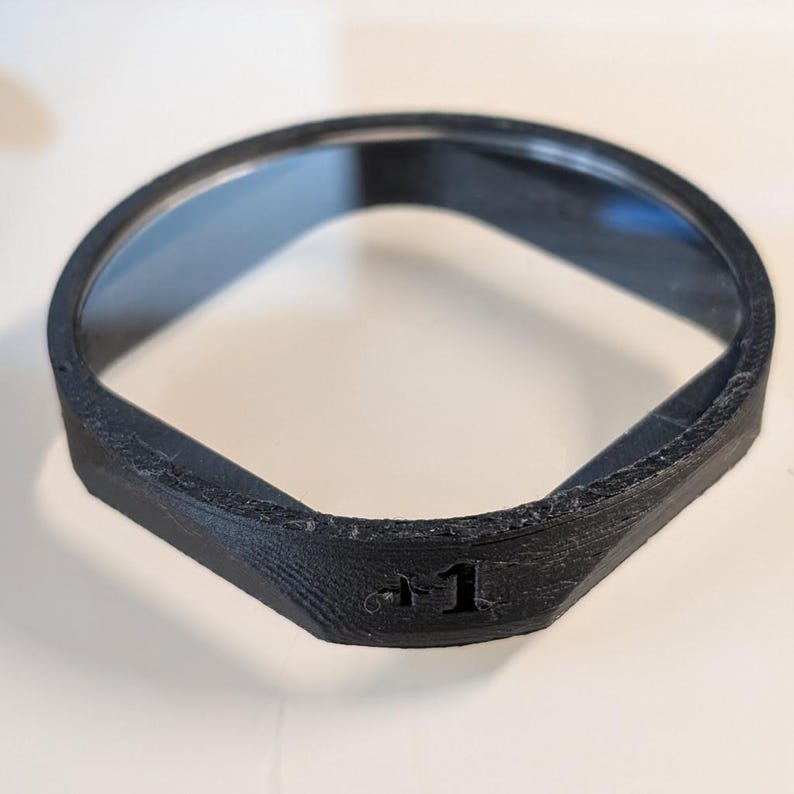 K&ouml;nnte beinhalten: Ein schwarzer, 3D-gedruckter Ring mit texturierter Oberfl&auml;che und einem gedrehten, nicht orientierbaren Schleifendesign. Der &auml;u&szlig;ere Rand des Bandes zeigt den gepr&auml;gten Text "+1", der auf der Vorderseite des Objekts mit hohler Mitte sichtbar ist.